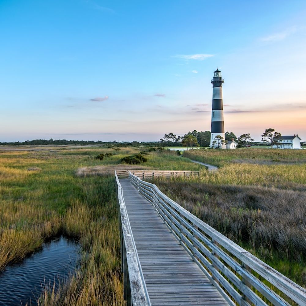 Lighthouse overlooking marshland and boardwalk at sunset, with a calm river and vibrant sky.
