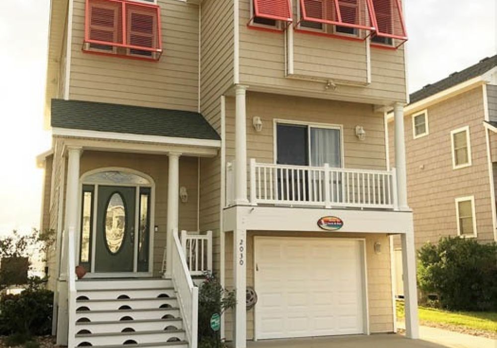 Modern beach house with red shutters and garage, featuring a welcoming front porch and green lawn.