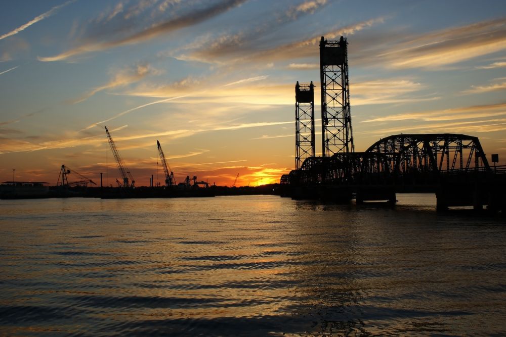 Sunset over a river with industrial cranes and a bridge silhouetted against the colorful sky.