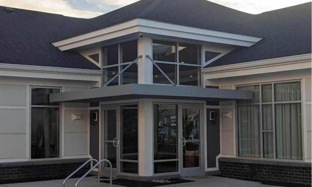 Modern building entrance with large glass doors, metal railing, and sloped roof under a partly cloudy sky.