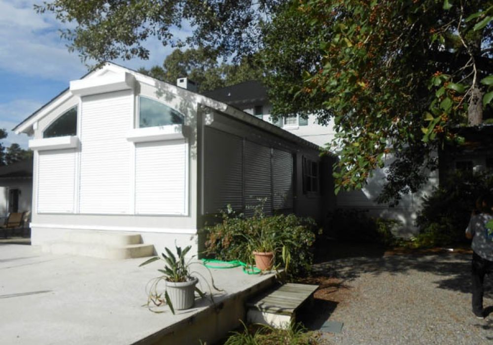 White house exterior with plants and shadows, showcasing energy-efficient roller shutters and sunny driveway.