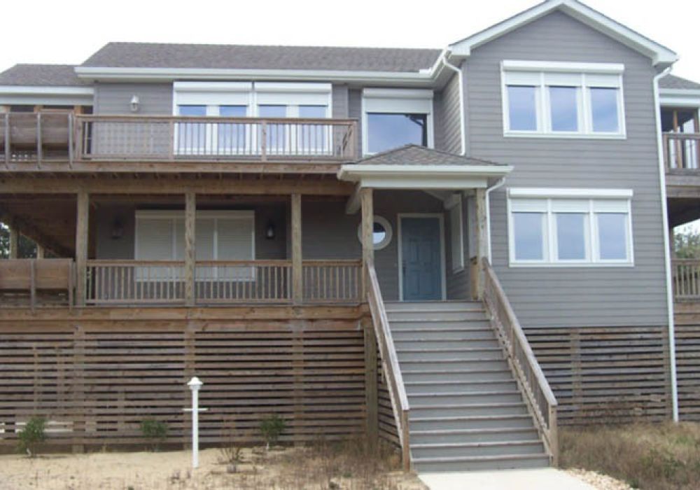 Gray two-story beach house with elevated wooden deck and staircase, surrounded by a sandy landscape and greenery.