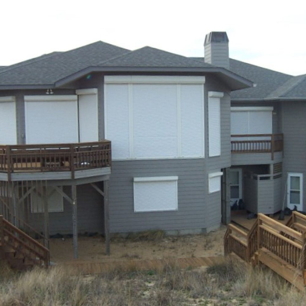 Gray beachfront house with wooden decks and sand dunes, featuring modern architecture and multiple staircases.