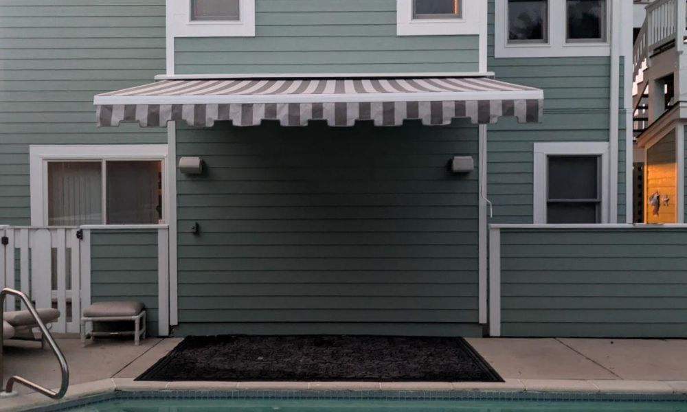 A turquoise pool with a diving board and a green house exterior with striped awning in the background.