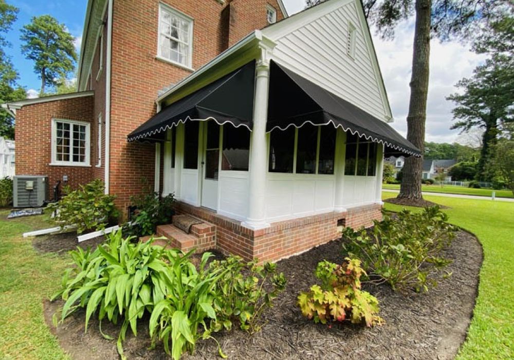Brick house with a black awning and lush garden, surrounded by trees and a green lawn on a sunny day.
