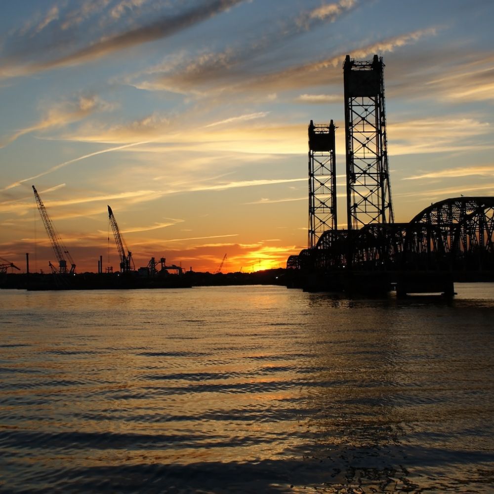 Sunset over a river with industrial cranes and a bridge silhouetted against the colorful sky.