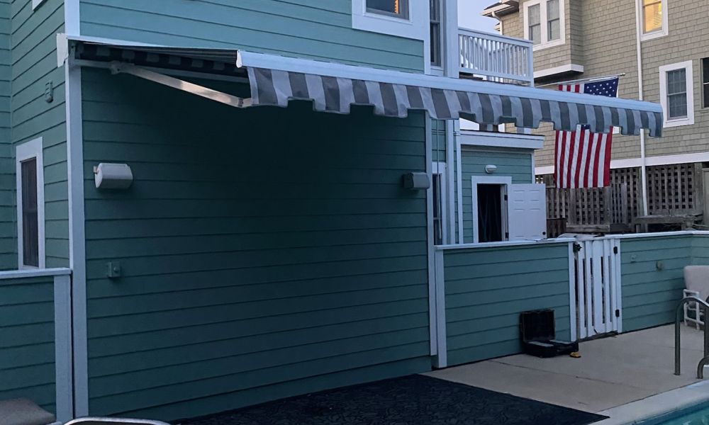 Modern blue house with a striped awning by a swimming pool, featuring a U.S. flag in the background.