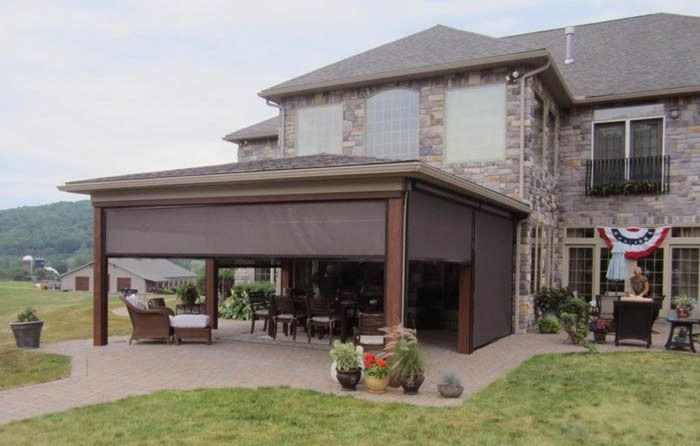 Large stone house with a furnished outdoor patio and a retractable awning, surrounded by a grassy yard and potted plants.