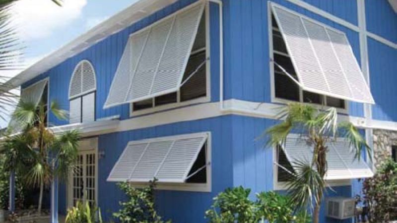 Blue house with open white window awnings, surrounded by tropical plants and trees under a clear sky.