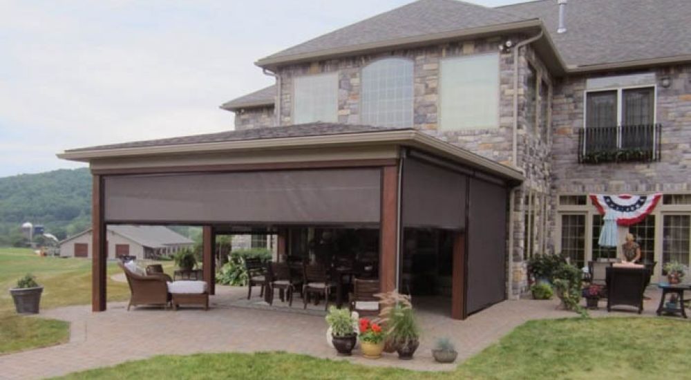 Large stone house with a furnished outdoor patio and a retractable awning, surrounded by a grassy yard and potted plants.