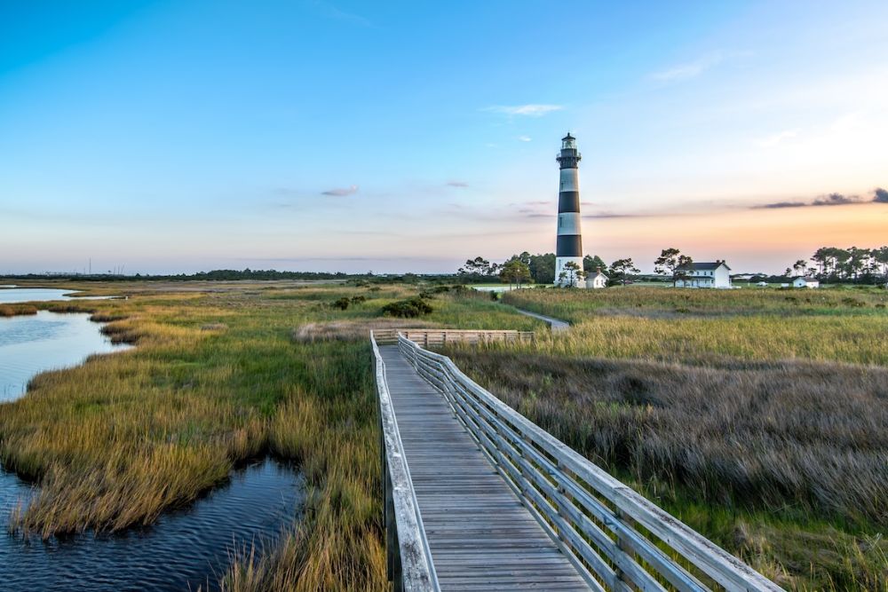 Lighthouse overlooking marshland and boardwalk at sunset, with a calm river and vibrant sky.