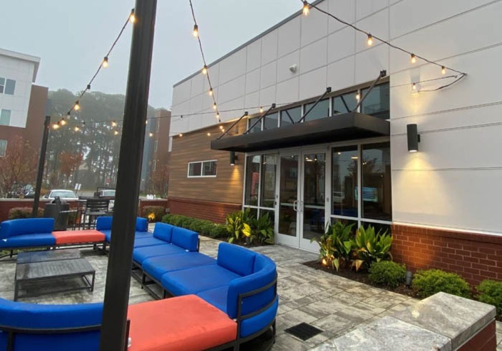 Modern patio with blue and orange seating outside a building, adorned with string lights and greenery.