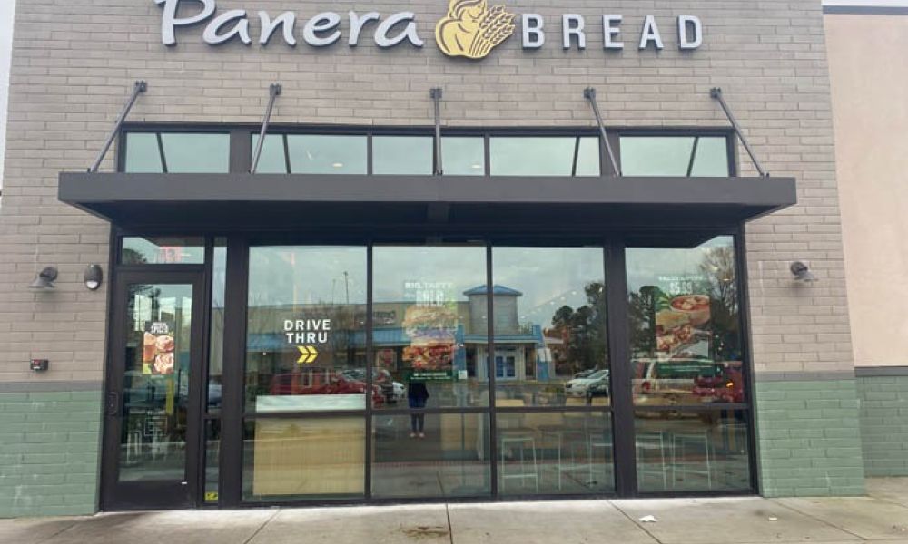 Exterior of a Panera Bread location with a drive-thru sign visible on the glass door.
