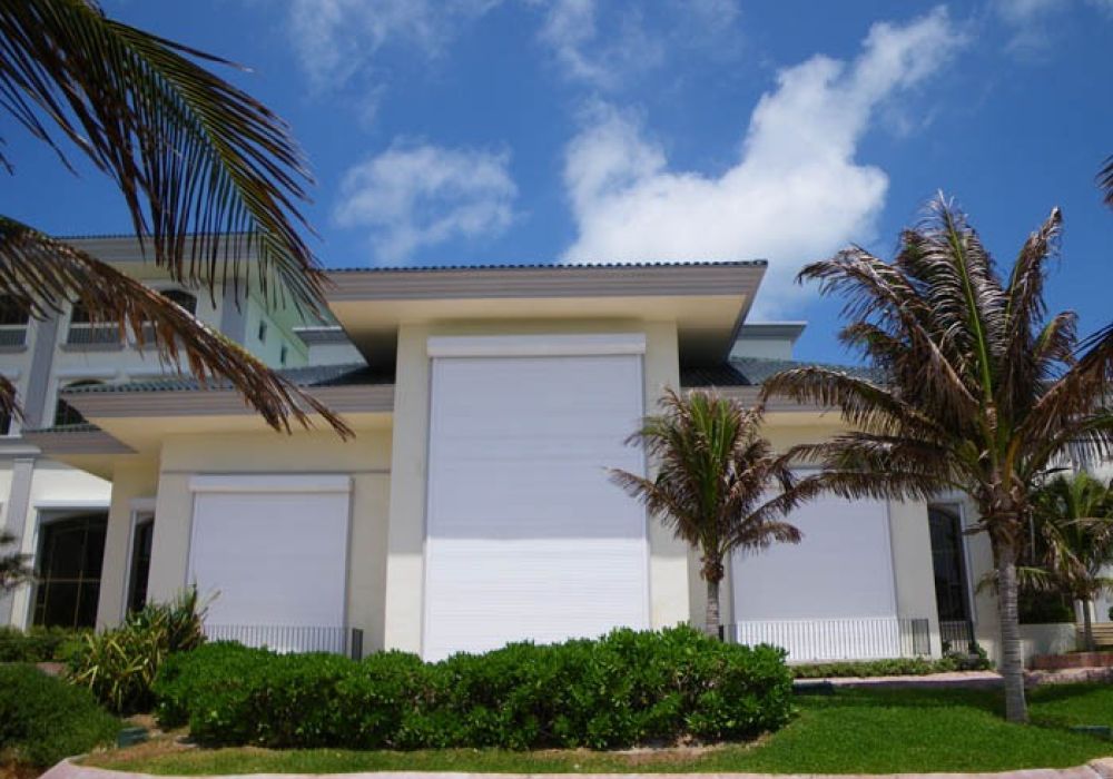 Modern coastal house with storm shutters and palm trees under a clear blue sky.