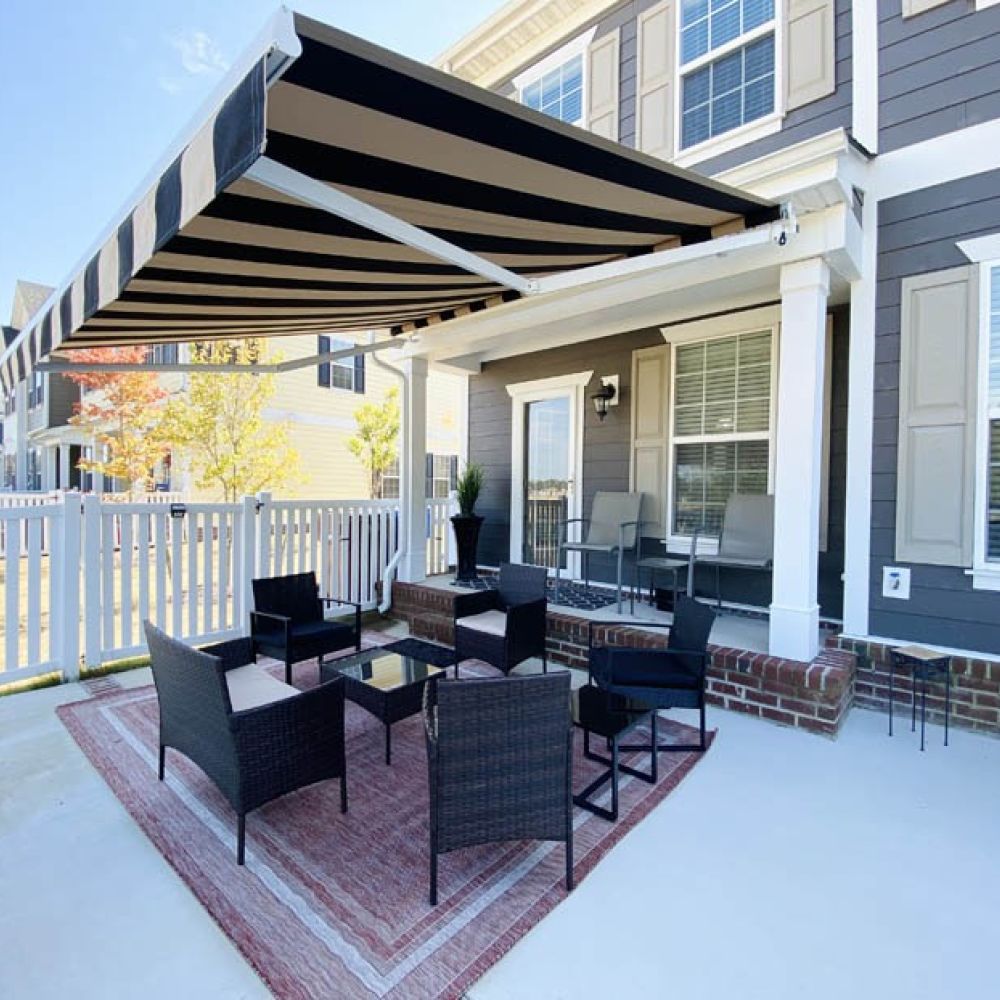 Modern patio with black outdoor furniture under striped retractable awning, adjacent to a grey house with white trim.