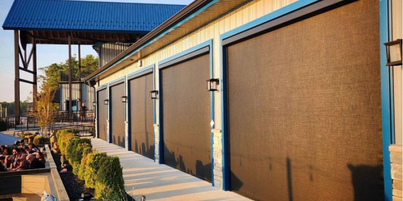 Modern building with outdoor screens and blue roof, surrounded by landscaping in bright sunlight.