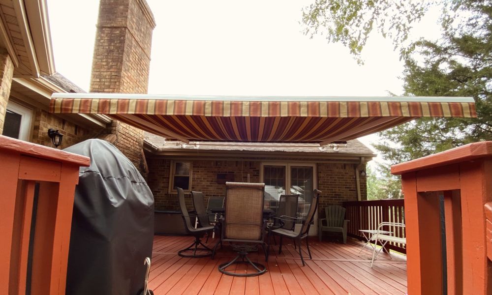 Striped patio awning over red deck with chairs and a grill in front of a brick house.