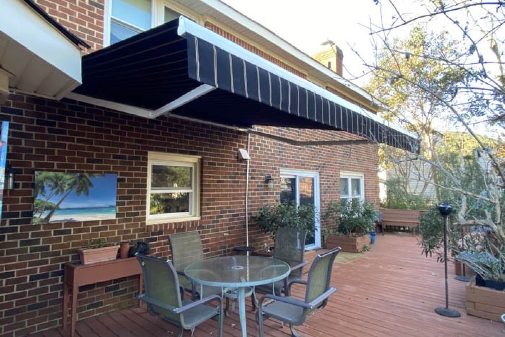Brick house patio with retractable awning, glass table, and chairs on wooden deck with greenery and outdoor decor.