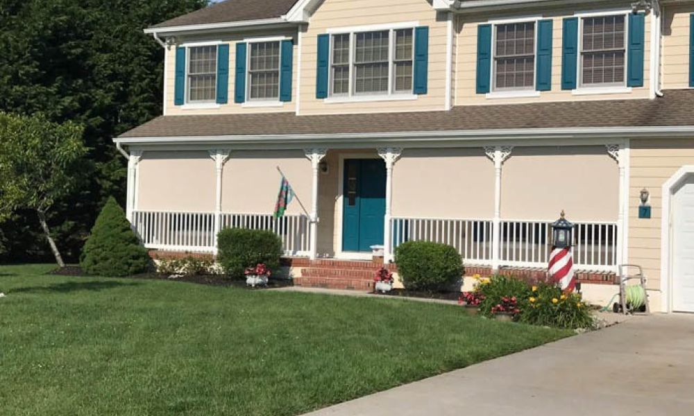 Suburban house with blue shutters, front porch, and manicured lawn under a clear sky.