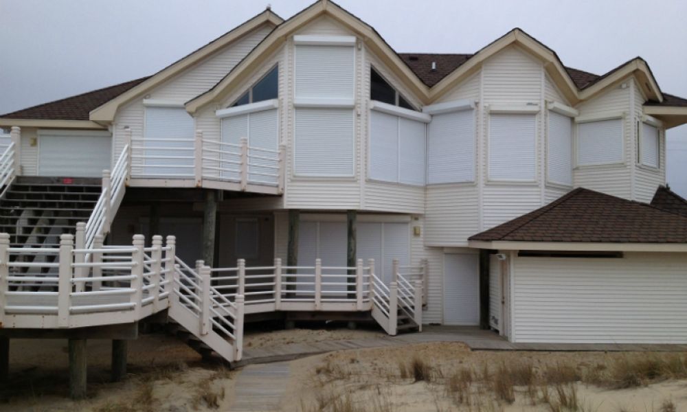 Modern beach house with white siding and large windows, situated on sand dunes under a cloudy sky.