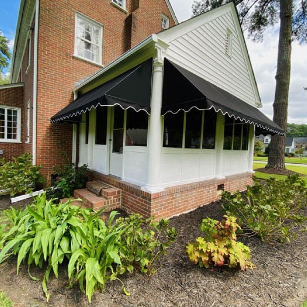 Brick house with a black awning and lush garden, surrounded by trees and a green lawn on a sunny day.