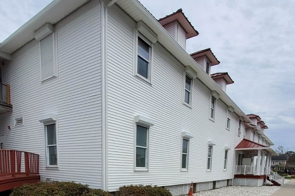 White two-story building with red roof accents, featuring multiple windows and a porch, under a cloudy sky.