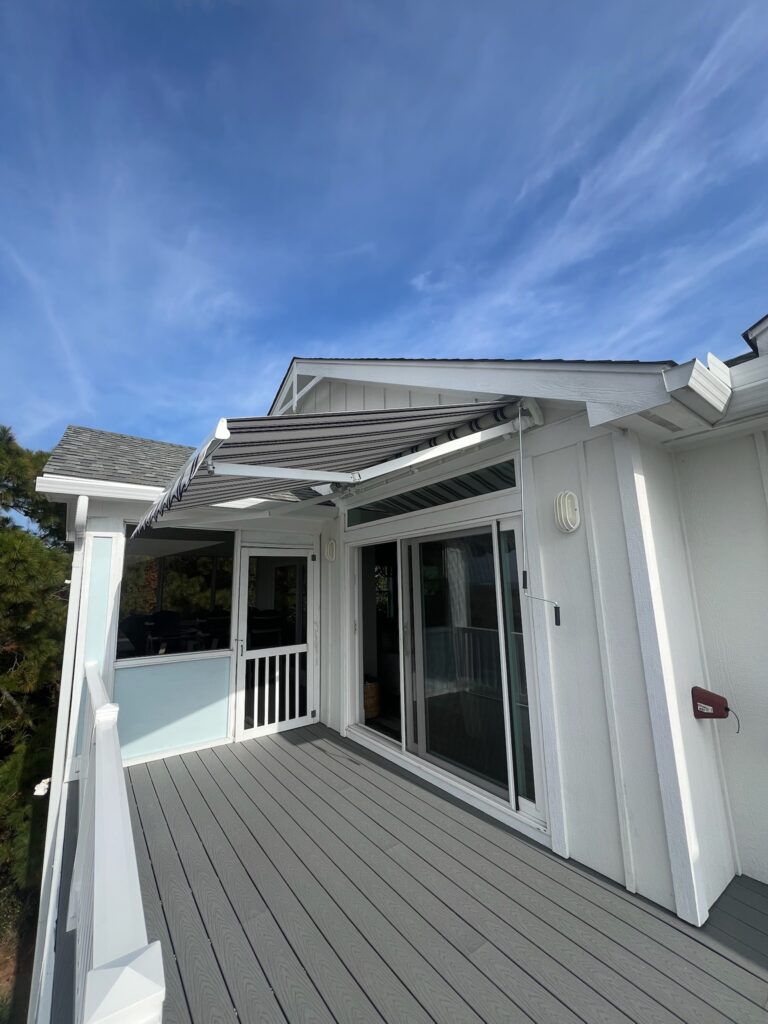 White house exterior with a gray wooden deck, sliding glass doors, and a striped retractable awning under a blue sky.