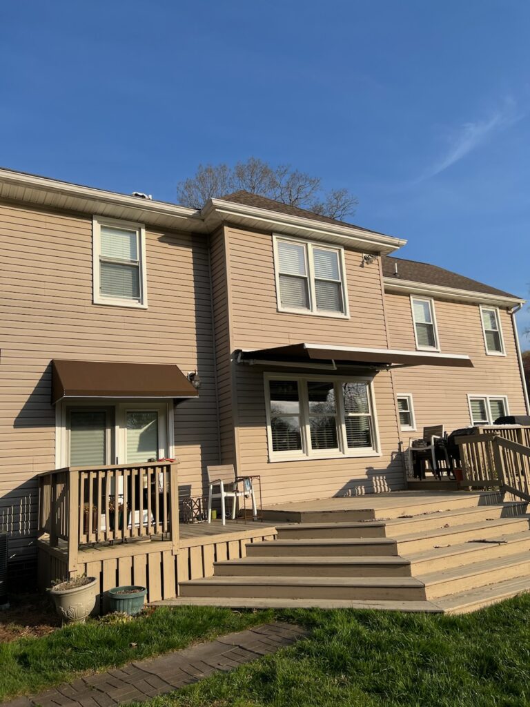 Two-story beige house with deck and brown awnings on a sunny day.
