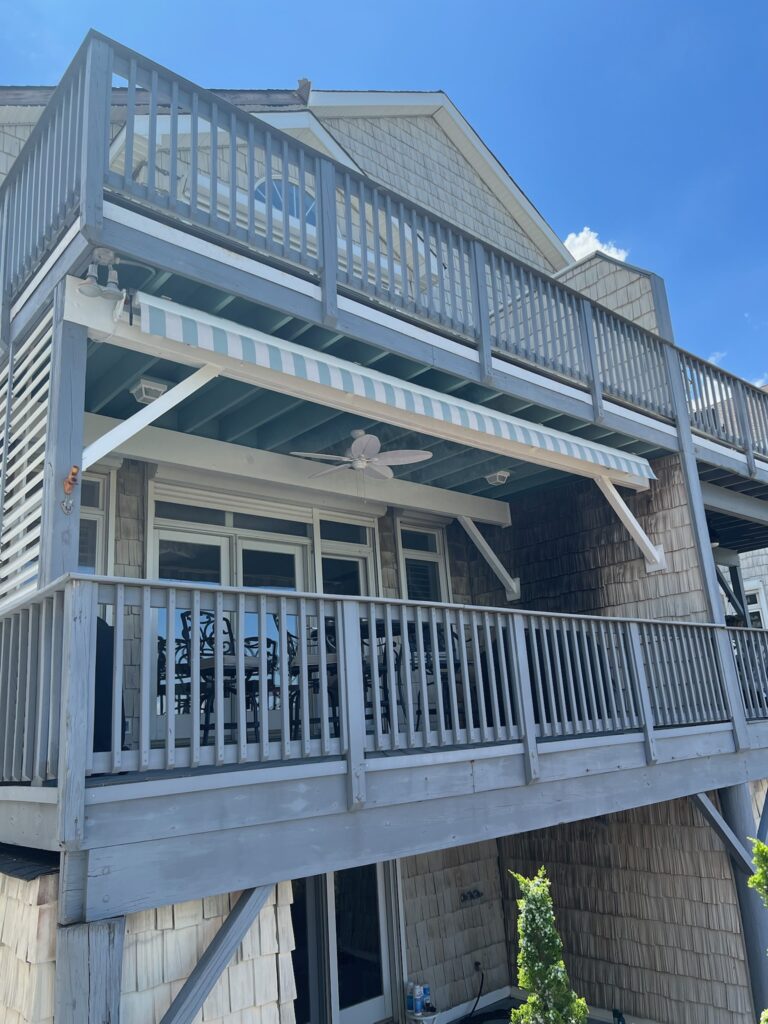 Two-story beach house with blue-gray wooden deck railings and a striped awning under a bright blue sky