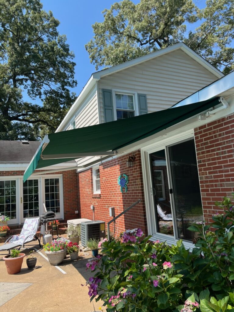 Brick house with a green awning, patio, potted plants, and flowers under a clear blue sky.