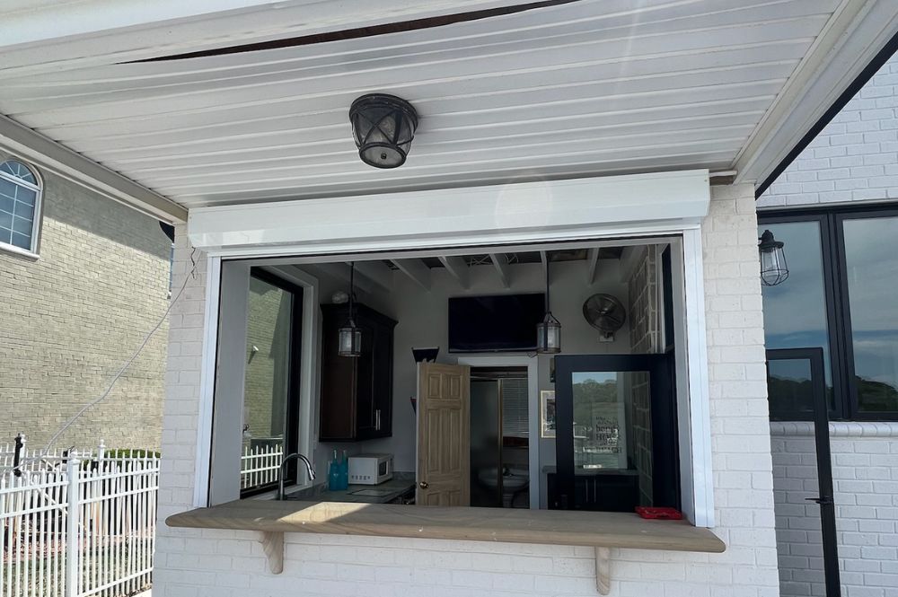 Outdoor bar window with wooden ledge, brick wall, and overhead lanterns, adjacent to a home with a white picket fence.